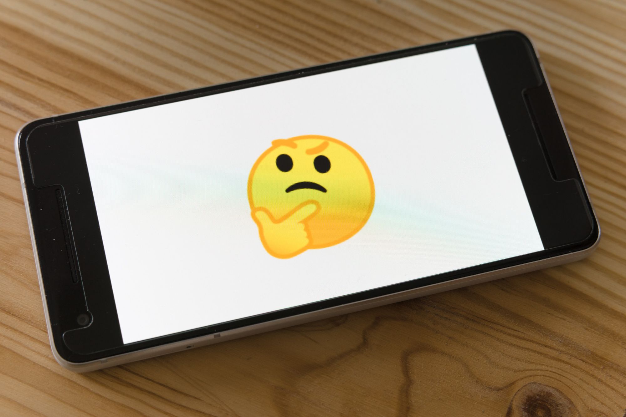 Phone on wooden background with a thinking emoji on the screen.