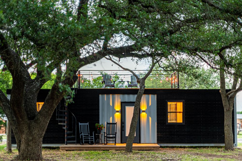 A tiny house made from a conex box with lawnchairs, trees, and front yard.